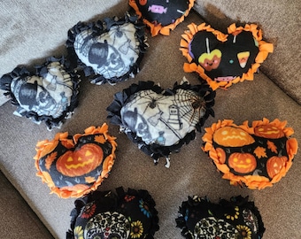 May include: A collection of eight heart-shaped fabric pillows featuring various Halloween-themed prints including skulls, pumpkins, spiderwebs, and candy. Each pillow is finished with a knotted fringe border in either solid orange or black.