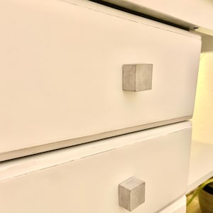 May include: White dresser with two drawers and concrete drawer pulls. The drawers are slightly open.