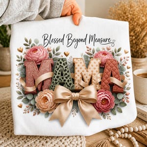 May include: A white t-shirt featuring the text Blessed Beyond Measure in black script above the word MAMA. The letters are decorated with glitter, leopard print, polka dots, and quilted textures, surrounded by pink peonies, green leaves, and a large beige bow.