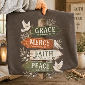 May include: A dark gray t-shirt featuring a graphic of a wooden signpost with four directional arrows. The signs read Grace Ephesians 2:8, Mercy Lamentations 3:22-23, Faith Hebrews 11:1, and Peace John 14:27, decorated with white doves and olive branches.