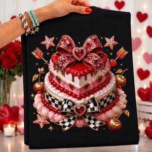 May include: A black t-shirt featuring a graphic of a multi-tiered heart-shaped cake decorated with red and white frosting, a large jeweled red bow, a black and white checkered ribbon, cherries, and stars, accented by golden arrows.