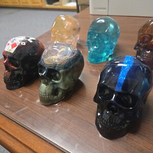 K&ouml;nnte beinhalten: Eine Sammlung dekorativer Totenkopf-Figuren in verschiedenen Farben und Designs. Die Sch&auml;del bestehen aus einem durchscheinenden Material, wobei einige aufwendige Muster und Akzente aufweisen. Zu den Farben geh&ouml;ren Schwarz, Blau, Orange und ein Totenkopf mit Spielkartenmotiven.