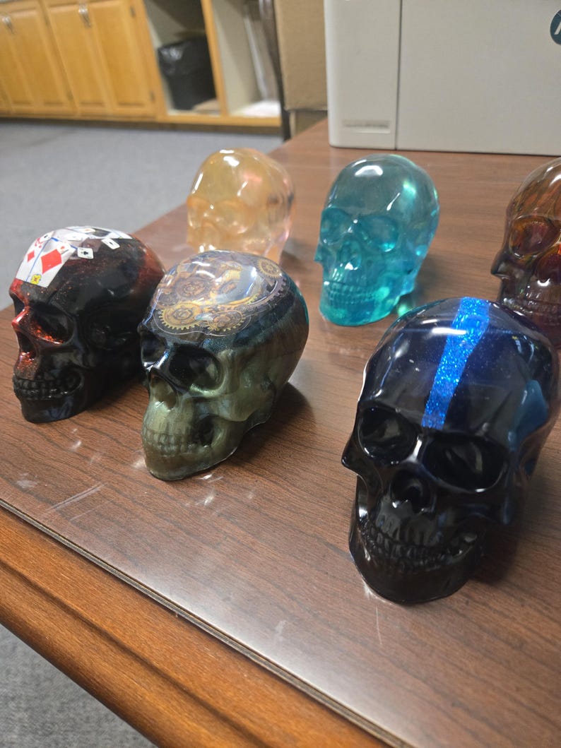 K&ouml;nnte beinhalten: Eine Sammlung dekorativer Totenkopf-Figuren in verschiedenen Farben und Designs. Die Sch&auml;del bestehen aus einem durchscheinenden Material, wobei einige aufwendige Muster und Akzente aufweisen. Zu den Farben geh&ouml;ren Schwarz, Blau, Orange und ein Totenkopf mit Spielkartenmotiven.