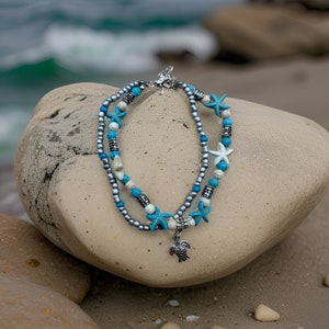 May include: A silver anklet with blue and white beads, starfish charms, and a silver turtle pendant. The anklet is displayed on a light brown rock.