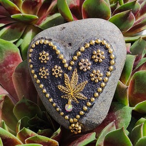 May include: A gray heart-shaped stone painted black with a gold marijuana leaf design and gold accents. The stone is surrounded by green and red succulents.