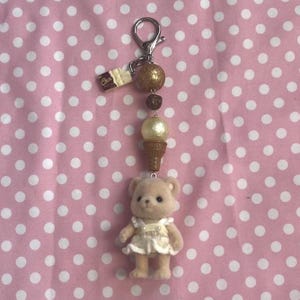 May include: A keychain featuring a small flocked bear figurine wearing a white dress, attached to a silver-tone metal clasp. The chain includes a brown ice cream cone charm, a cream-colored bead, a dark brown bead, a gold glitter bead, and a small chocolate bar charm.