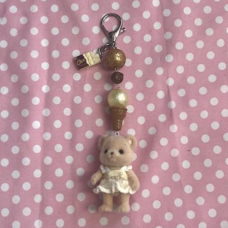 May include: A keychain featuring a small flocked bear figurine wearing a white dress, attached to a silver-tone metal clasp. The chain includes a brown ice cream cone charm, a cream-colored bead, a dark brown bead, a gold glitter bead, and a small chocolate bar charm.