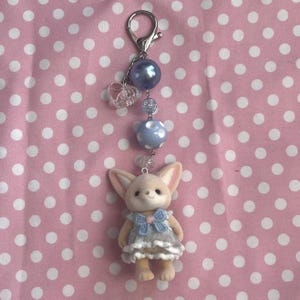 May include: A keychain featuring a small, beige, flocked animal figurine wearing a blue and white dress with a bow. The charm is attached to a silver-toned lobster clasp by a chain of beads, including a large blue pearl, a clear heart, and a blue polka-dot bead.