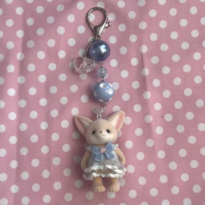 May include: A keychain featuring a small, beige, flocked animal figurine wearing a blue and white dress with a bow. The charm is attached to a silver-toned lobster clasp by a chain of beads, including a large blue pearl, a clear heart, and a blue polka-dot bead.