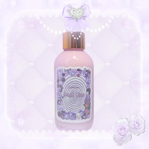 Puede incluir: Una botella rosa con tapa dorada y una etiqueta que dice "fresh plum". La etiqueta presenta un dise&ntilde;o de remolino y varias ilustraciones moradas. Un lazo morado con un coraz&oacute;n plateado se encuentra encima de la botella. El fondo es morado suave.