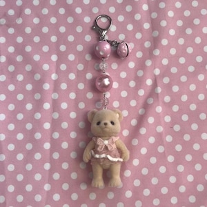 May include: A keychain featuring a small, beige, flocked bear figurine wearing a pink dress with a bow. The bear is attached to a silver-toned lobster clasp by a chain of beads, including a large pink polka-dot bead, a clear bead, and a pink pearl.