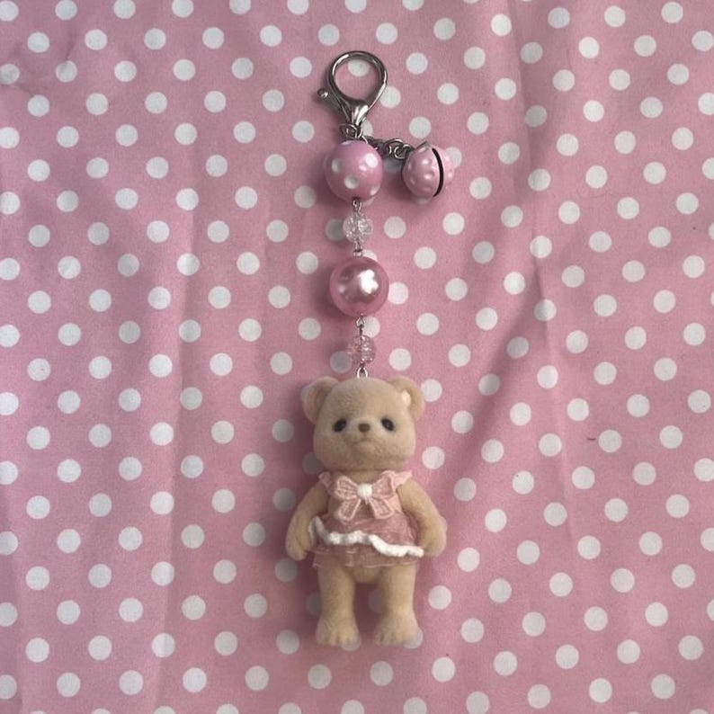 May include: A keychain featuring a small, beige, flocked bear figurine wearing a pink dress with a bow. The bear is attached to a silver-toned lobster clasp by a chain of beads, including a large pink polka-dot bead, a clear bead, and a pink pearl.