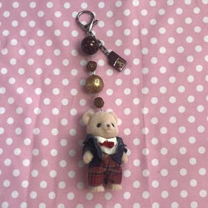 May include: A keychain featuring a small flocked bear figurine wearing a navy blue jacket, white shirt with a red bow tie, and red plaid trousers. The bear hangs from a silver-tone metal clasp with brown beads and a small rectangular charm.