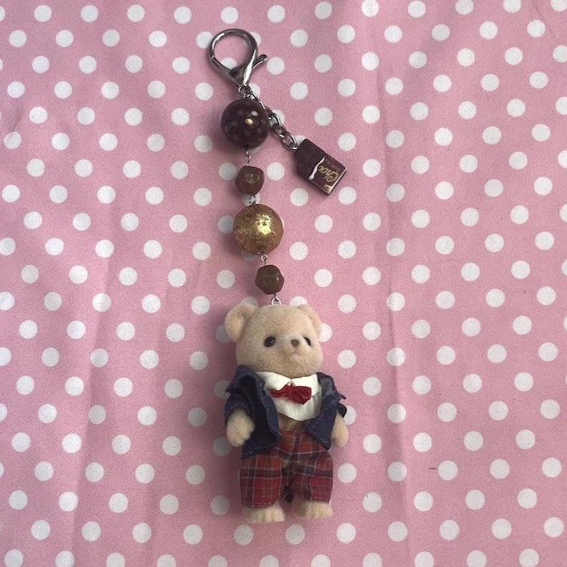 May include: A keychain featuring a small flocked bear figurine wearing a navy blue jacket, white shirt with a red bow tie, and red plaid trousers. The bear hangs from a silver-tone metal clasp with brown beads and a small rectangular charm.