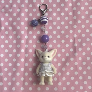 May include: A keychain featuring a small, cream-colored fennec fox figurine wearing a white ruffled dress. The fox is attached to a silver-toned lobster clasp by a chain of beads, including a purple striped bead, a solid purple bead, and clear beads.