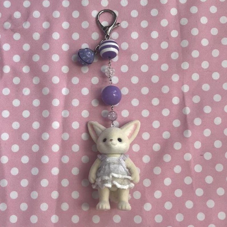 May include: A keychain featuring a small, cream-colored fennec fox figurine wearing a white ruffled dress. The fox is attached to a silver-toned lobster clasp by a chain of beads, including a purple striped bead, a solid purple bead, and clear beads.