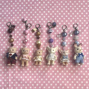 May include: Six miniature animal figurines attached to silver-toned metal keychains with beaded accents. The figures include three bears, two fennec foxes, and one rabbit, each wearing detailed fabric clothing in various colors and patterns.