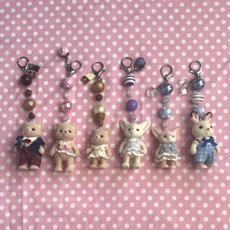 May include: Six miniature animal figurines attached to silver-toned metal keychains with beaded accents. The figures include three bears, two fennec foxes, and one rabbit, each wearing detailed fabric clothing in various colors and patterns.