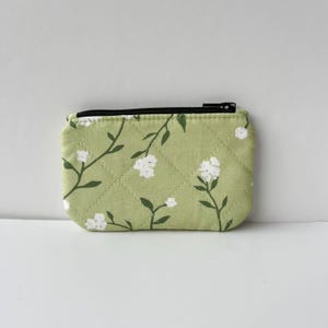 May include: A small, quilted coin purse with a pale green background and a floral pattern of white flowers and dark green stems. The purse has a black zip closure and a rectangular shape.