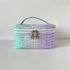 May include: A small, quilted cosmetic bag with a handle. The bag features a repeating pattern of white, teal, gray, and purple floral designs. It has a white zipper and two silver zipper pulls. The handle is teal and purple.