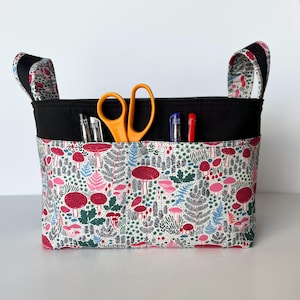 Puede incluir: Una cesta de almacenamiento de tela con la parte superior y asas negras. La cesta presenta un colorido estampado de setas rojas, helechos rosas y follaje verde sobre un fondo blanco. En el interior hay bolígrafos y tijeras naranjas.