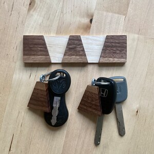 May include: A wooden key rack with a geometric pattern in shades of brown and white. Two keychains with keys are hanging on the rack. One keychain has a brown wooden charm and a black car key. The other keychain has a brown wooden charm and a silver car key.