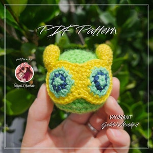 May include: A green and yellow crocheted amigurumi toy of Gekko from the video game Valorant. The toy has a yellow mask with green and blue eyes. The text "VALORANT Gekko's Moshpit" is visible below the toy.