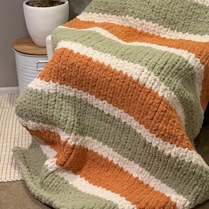 May include: A thick, striped blanket in shades of orange, sage green, and white. The blanket is displayed over a chair, highlighting its soft texture and colour scheme. This cosy throw blanket is ideal for adding warmth and style to any living area.