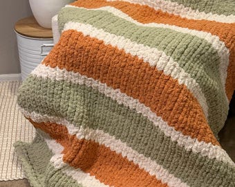 May include: A thick, striped blanket in shades of orange, sage green, and white. The blanket is displayed over a chair, highlighting its soft texture and colour scheme. This cosy throw blanket is ideal for adding warmth and style to any living area.