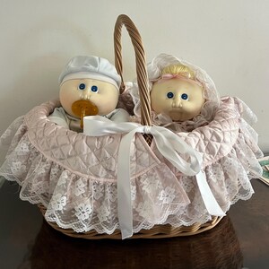 Puede incluir: Una cesta de mimbre con asa contiene dos muñecas. Las muñecas tienen ojos azules y llevan sombreros. La cesta está forrada con tela acolchada rosa y ribete de encaje blanco. Una cinta blanca está atada al frente.