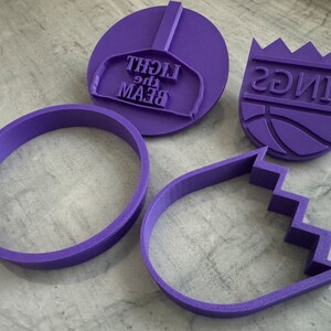 May include: Four purple plastic cookie cutters. One is a circle, one is an oval with a jagged edge, one is a basketball with the words "KINGS" on it, and one is a baseball cap with the words "BEAM the LIGHT" on it.