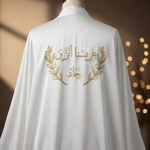 May include: White robe with gold embroidered Arabic calligraphy and laurel wreath design. The robe is draped over a mannequin, showcasing the back of the garment. The text is in gold thread.