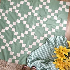 May include: A square quilt featuring a repeating checkerboard pattern in sage green and off-white fabric squares. The quilt is displayed alongside a green and white gingham fabric cloth and a bouquet of bright yellow lily flowers.