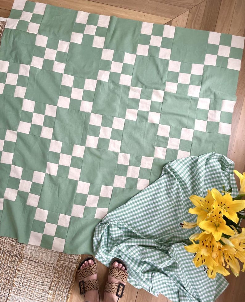 May include: A square quilt featuring a repeating checkerboard pattern in sage green and off-white fabric squares. The quilt is displayed alongside a green and white gingham fabric cloth and a bouquet of bright yellow lily flowers.