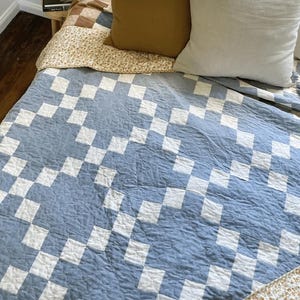 May include: A quilt featuring a repeating geometric pattern of light blue and white squares arranged in a chain-like design. The fabric has a visible quilted texture across the surface, covering the bed alongside a solid mustard and white pillow.