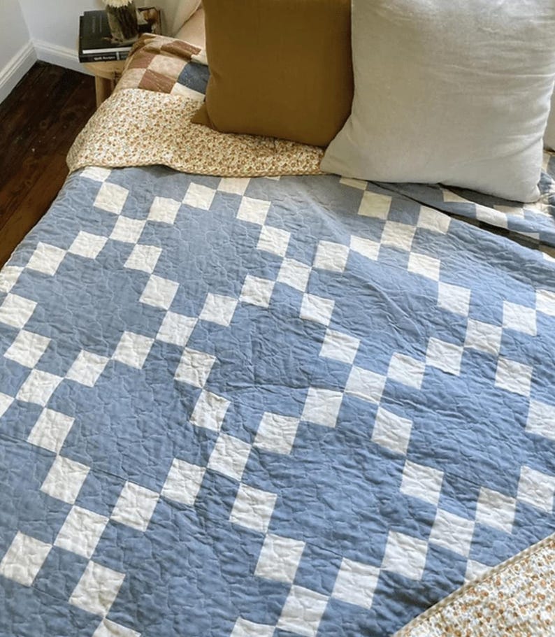 May include: A quilt featuring a repeating geometric pattern of light blue and white squares arranged in a chain-like design. The fabric has a visible quilted texture across the surface, covering the bed alongside a solid mustard and white pillow.