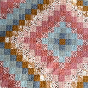 May include: A patchwork quilt with a geometric design. The quilt features squares in shades of pink, blue, brown, and white. The squares are arranged in a grid pattern, creating a visually appealing and textured surface.