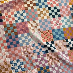 May include: A patchwork quilt with a variety of square patterns in shades of pink, blue, brown, and yellow. Some squares feature floral and bird designs, while others have checkered or solid patterns. The quilt is folded, showing its layered construction.