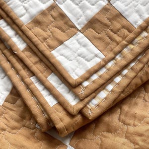 May include: A quilted blanket featuring a repeating checkerboard pattern in tan and white. The fabric has a visible texture with stitched detailing throughout, including white thread accents along the edges and a finished bound border.