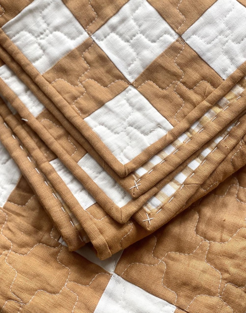 May include: A quilted blanket featuring a repeating checkerboard pattern in tan and white. The fabric has a visible texture with stitched detailing throughout, including white thread accents along the edges and a finished bound border.
