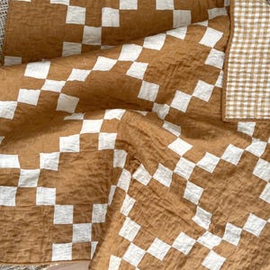May include: A quilted blanket featuring a large-scale brown and white checkerboard pattern on one side and a smaller brown and white gingham check pattern on the reverse. The fabric has a crinkled, textured surface throughout the entire piece.