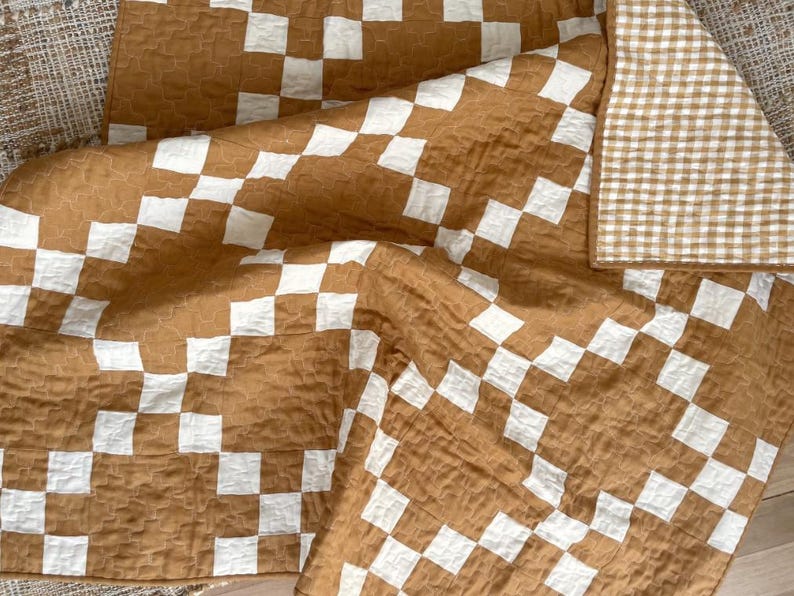 May include: A quilted blanket featuring a large-scale brown and white checkerboard pattern on one side and a smaller brown and white gingham check pattern on the reverse. The fabric has a crinkled, textured surface throughout the entire piece.