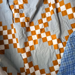 May include: A quilt featuring a repeating checkerboard pattern in mustard yellow, white, and light blue fabric squares. The textile displays a grid-like arrangement of these colored blocks, with a section of blue plaid fabric visible at the bottom right.