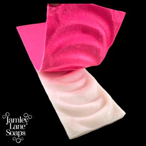 May include: Two bars of handmade soap, one bright pink and one pale pink, are displayed against a black background. The top bar is folded over the bottom bar, revealing a swirl pattern. The logo "Jamley Lane Soaps" is in the lower left corner.