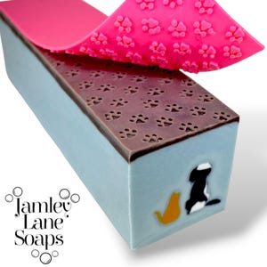May include: A rectangular bar of soap with a light blue base, topped with a brown layer featuring paw prints. A pink sheet with paw prints is partially peeled back. The side of the soap features a black and white dog and an orange cat. The text "Jamley Lane Soaps" is visible.