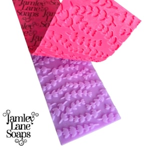 May include: A rectangular silicone texture mat featuring a raised lavender sprig pattern. The mat is light purple with a vibrant pink section folded over the top. The text Jamley Lane Soaps is printed in black on the upper left corner of the mat.