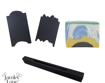 May include: Three black silicone soap moulds, one with a straight edge, one with a wavy edge, and one long rectangular mould with a groove in the centre. The moulds are on a white background.