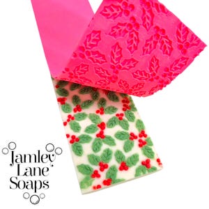 Può includere: Una saponetta decorata con foglie di agrifoglio verdi e bacche rosse su sfondo bianco. Un foglio rosa con un motivo a foglie di agrifoglio è parzialmente staccato, rivelando il design del sapone. Il testo "Jamley Lane Soaps" è visibile.