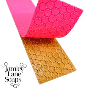Puede incluir: Primer plano de un molde de jabón rosa y dorado con un patrón de panal. La sección rosa se levanta para revelar la sección dorada debajo. Las palabras "Jamley Lane Soaps" están en la esquina inferior izquierda.