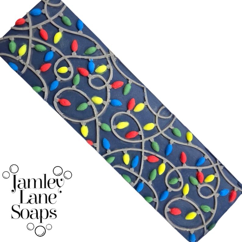 May include: A dark blue rectangular soap mold featuring a string of colorful Christmas lights. The lights are red, yellow, green, and blue. The mold is made of a flexible material, and the design is raised. The brand name "Jamley Lane Soaps" is in the lower left corner.